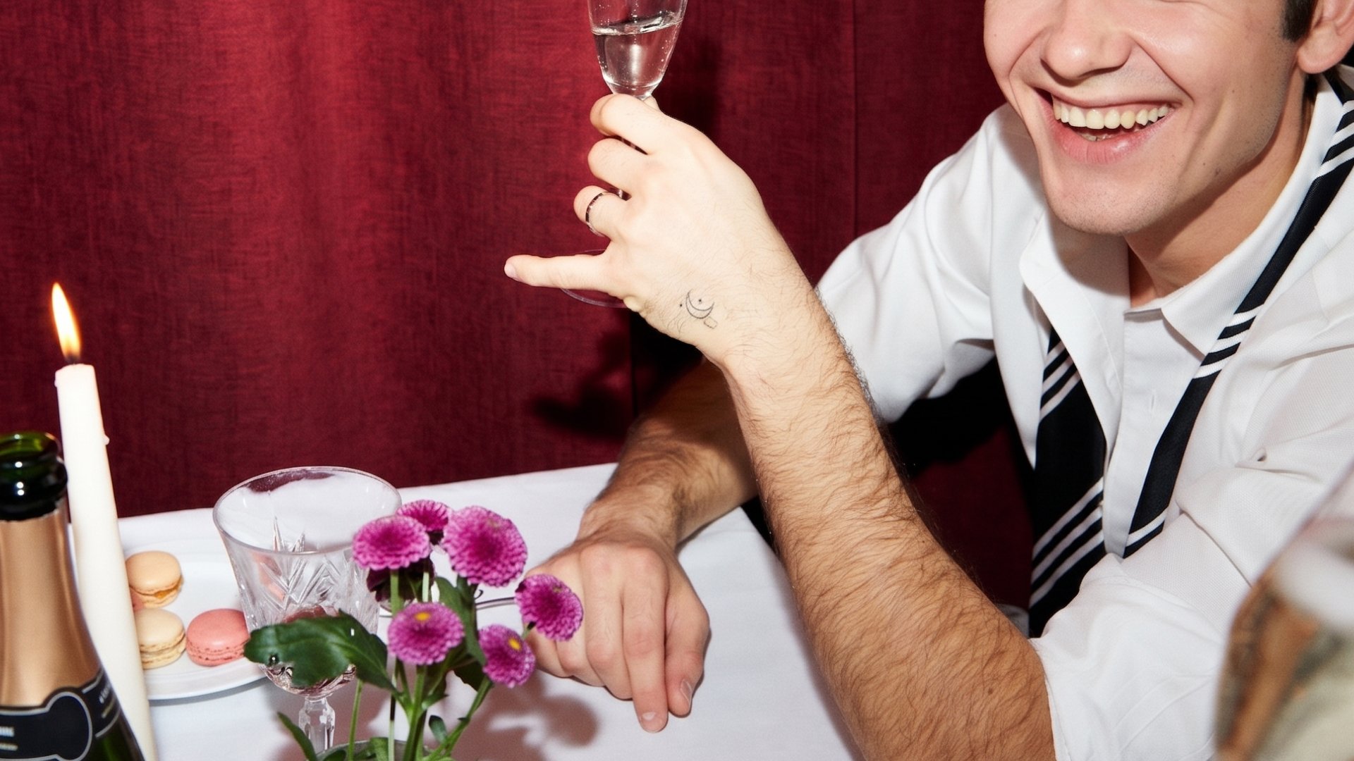 Smiling man in white shirt with hand tattoo holding a wine glass at a table with candle, flowers, and macarons.